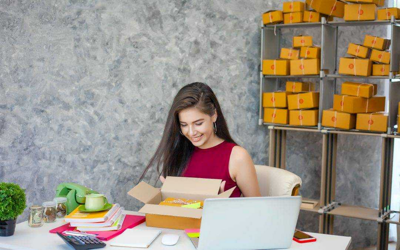 A woman is sitting at a desk, smiling while opening a cardboard box with more boxes stacked on shelves behind her.