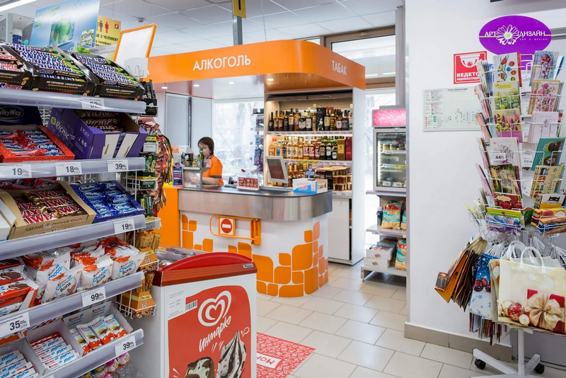 A convenience store interior showing a cashier at the counter, shelves stocked with various items, and signage in Cyrillic.