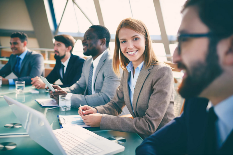 A group of business professionals sits at a table during a meeting, with one woman smiling at the camera.