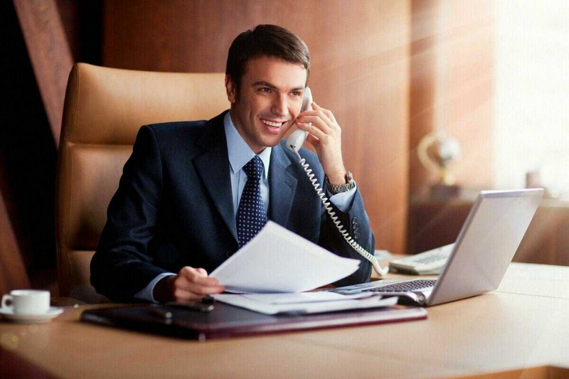 A man in a suit is smiling and talking on the phone while holding papers and sitting at a desk with a laptop.